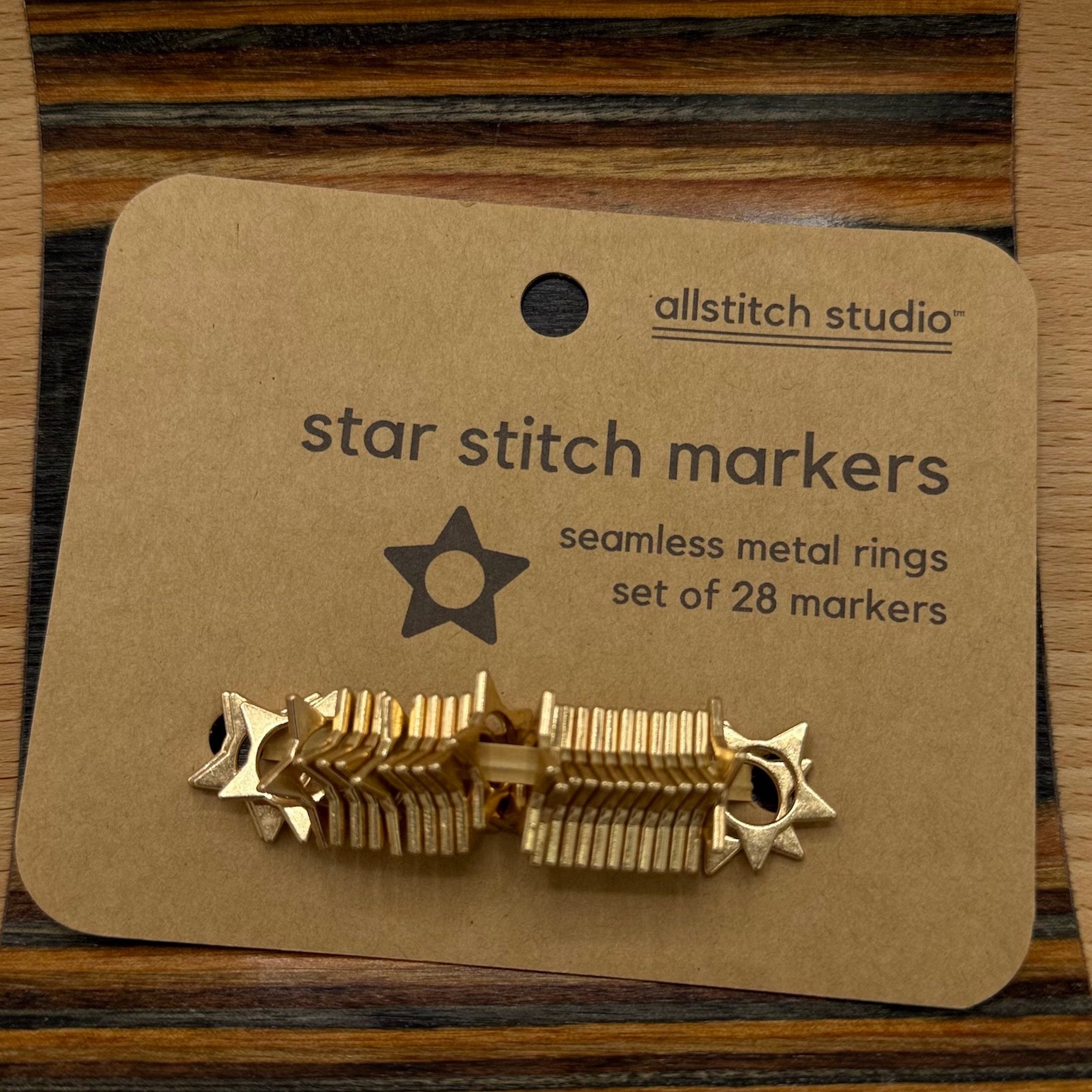 Gold star stitch markers on a brown card with 'allstitch studio' branding on a wooden surface.