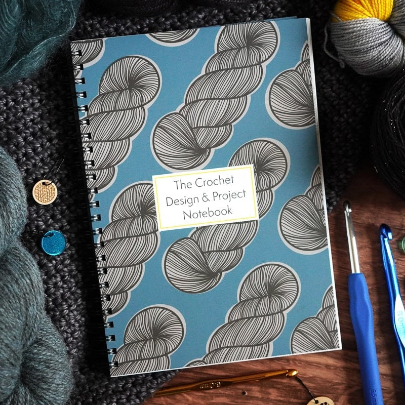 Notebook titled 'The Crochet Design & Project Notebook' with yarn and crochet tools on a wooden surface
