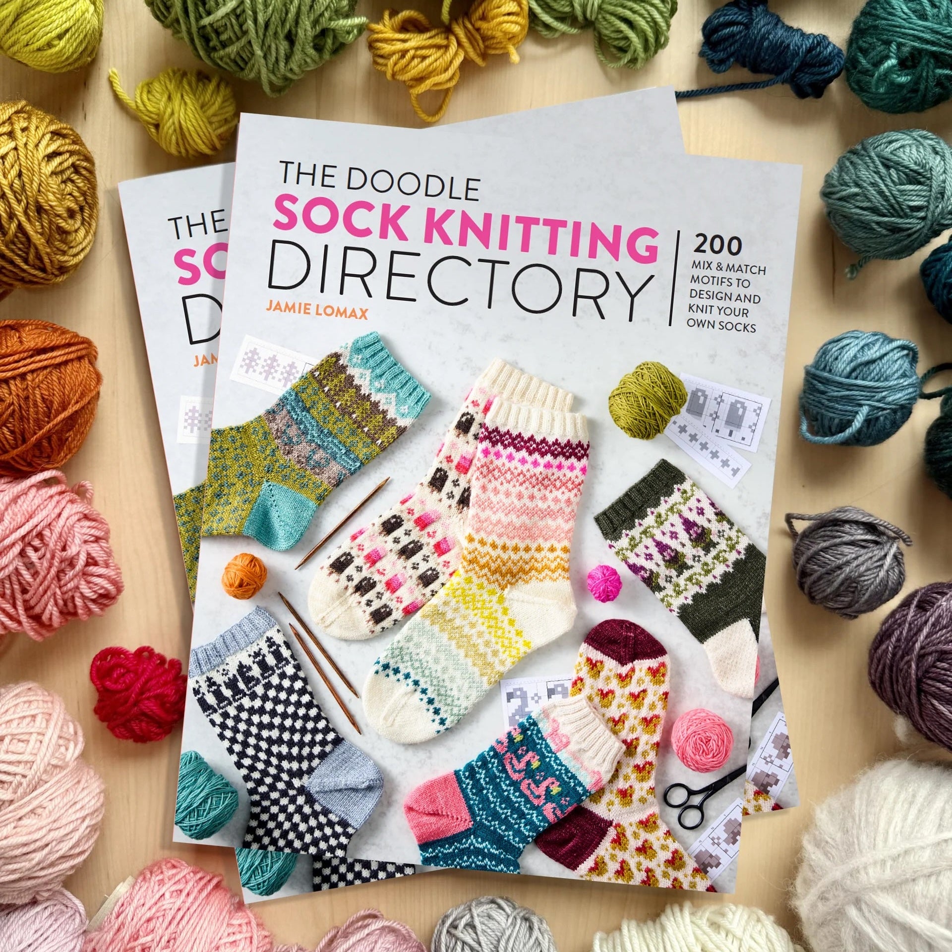 The Doodle Sock Knitting Directory book by Jamie Lomax, styled in a flat lay with various colorful balls of yarn surrounding it on a light wood background.
