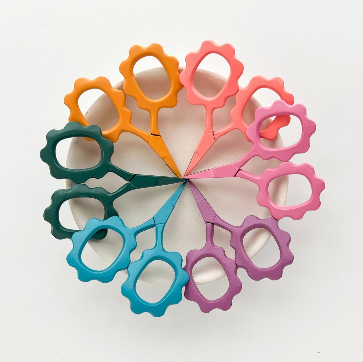 Six small flower-shaped scissors arranged in a circular fan pattern on a white background, featuring handles in orange, coral pink, magenta, purple, teal blue, and dark green.