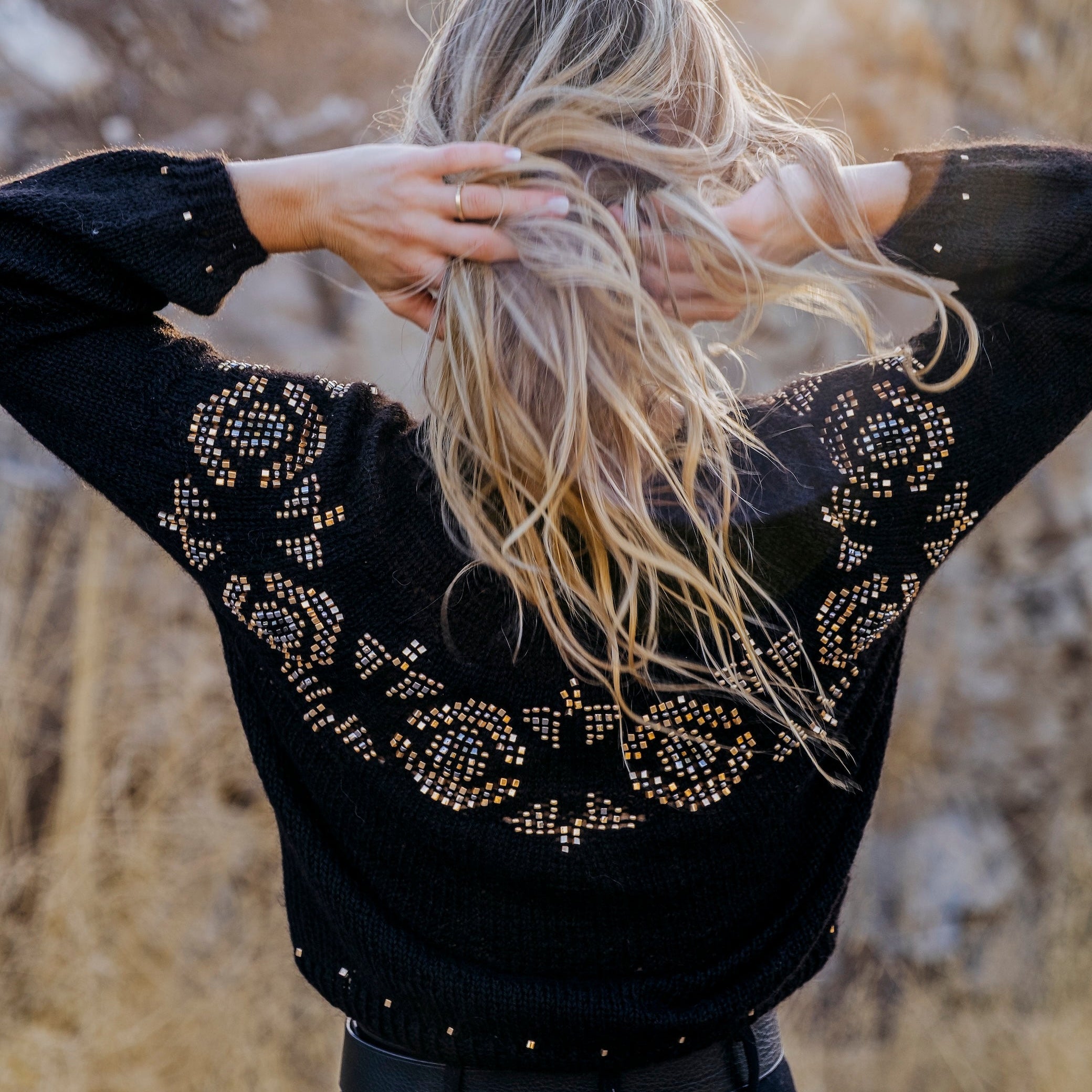 A woman viewed from behind wearing a black knit sweater with intricate gold rhinestone patterns across the shoulders and upper back, paired with black pants, set against a blurred natural background of dry grass and rocks.