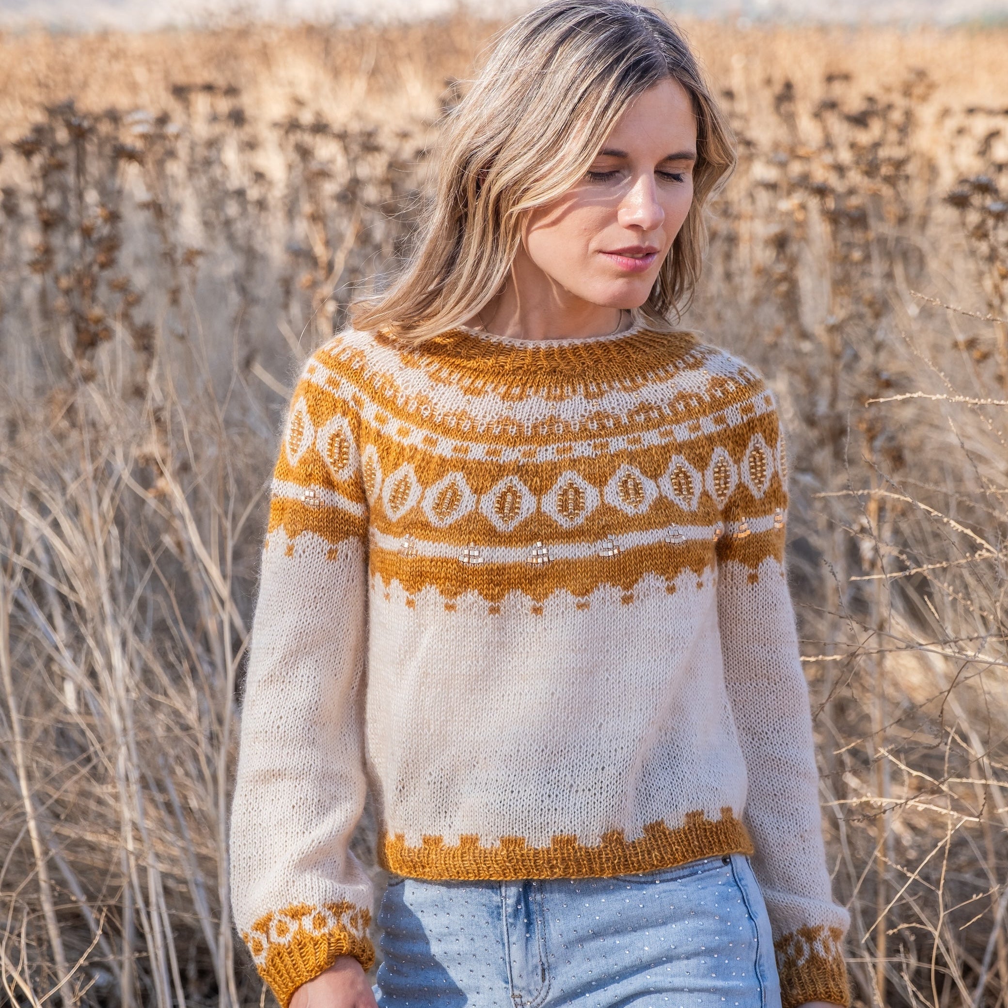 A woman wearing a cream and mustard yellow Fair Isle knit sweater with light blue jeans, standing in a field of dry grass.