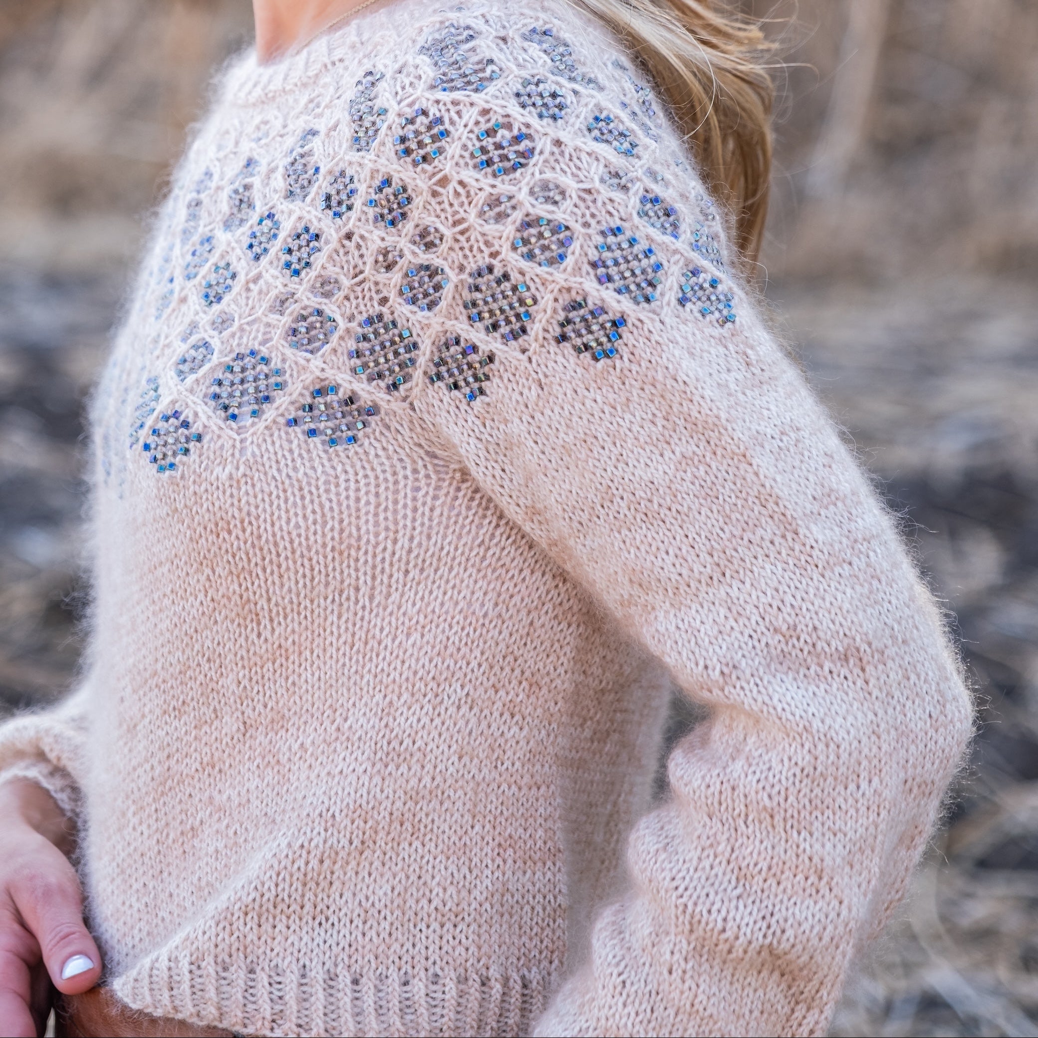 A woman wearing a fuzzy knit sweater in a natural outdoor setting. The sweater is a light beige color with a textured, geometric lace-like pattern in a muted blue-gray on the yoke and shoulders.