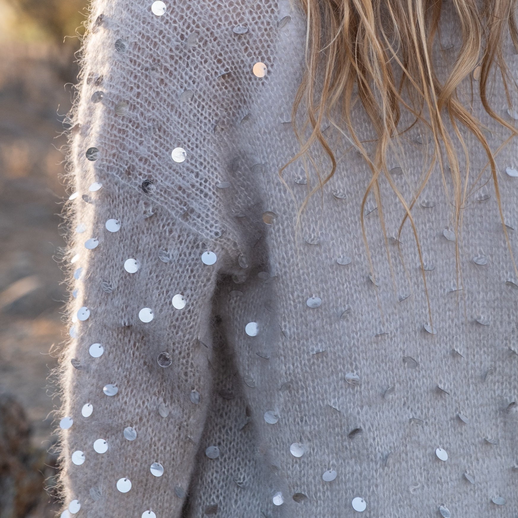 Close-up of a person wearing a fuzzy light gray knit sweater covered with scattered silver sequins, with long blonde hair visible against a softly blurred outdoor background.