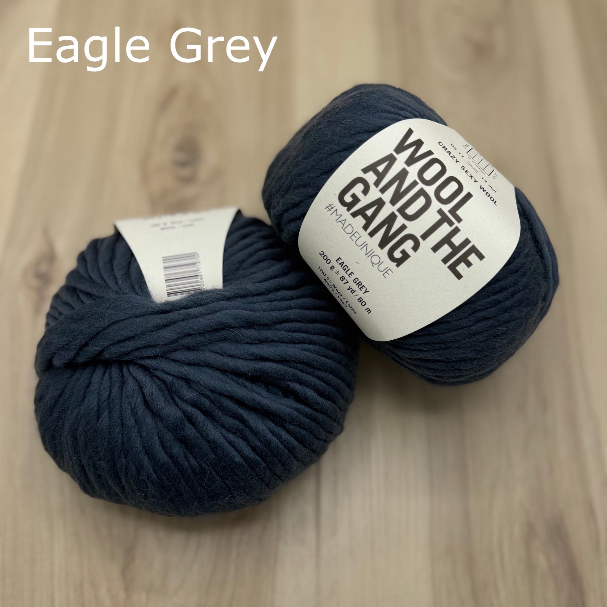 Two balls of super-bulky dark grey-blue yarn with 'Wool and the Gang' label on a wooden surface, labeled 'Eagle Grey'.