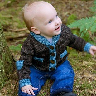 A baby sitting on mossy ground wearing a brown and blue knitted cardigan